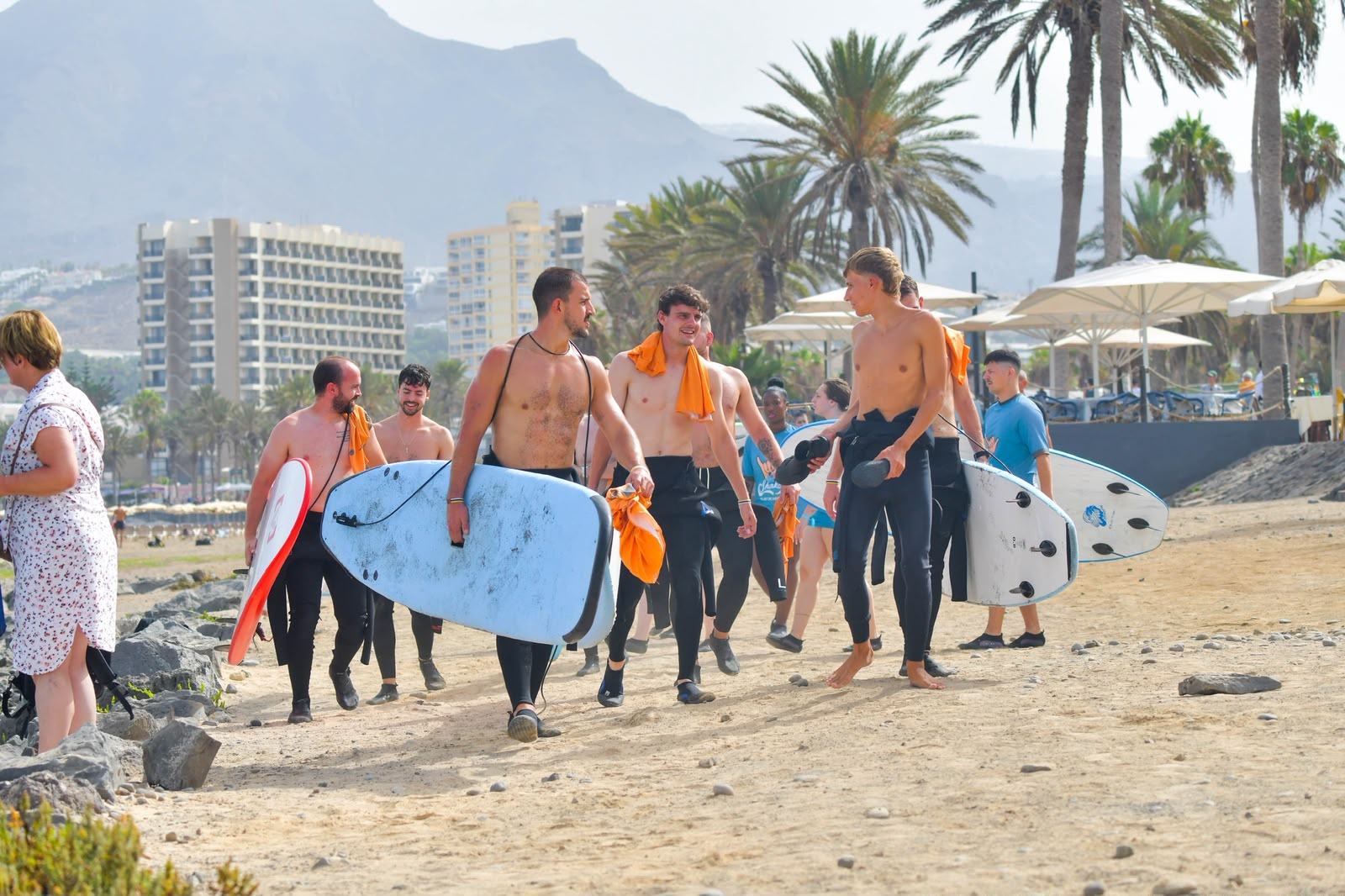 Schüler der Klasse Kontraola spazieren mit ihren Surfbrettern durch Playa de las Américas, im Hintergrund sind die Hotels von Las Américas zu sehen